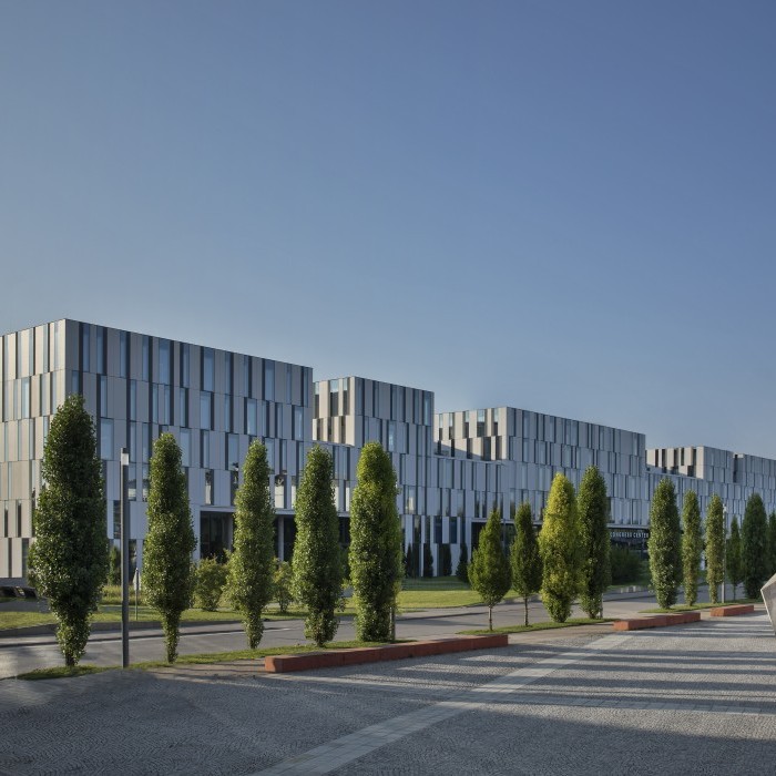 Moderne Bürogebäude mit geometrischer Fassade, gesäumt von gepflegten Bäumen und einer klaren, gepflasterten Promenade unter blauem Himmel