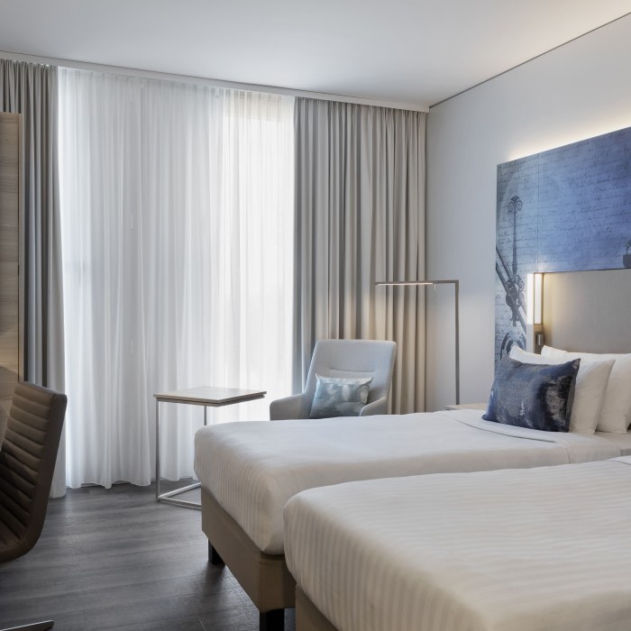 Helles Hotelzimmer mit zwei Einzelbetten, modernem Schreibtisch, Sitzbereich und dekorativen blauen Akzenten vor bodentiefen Vorhängen in einem Hotel nahe dem Flughafen München.