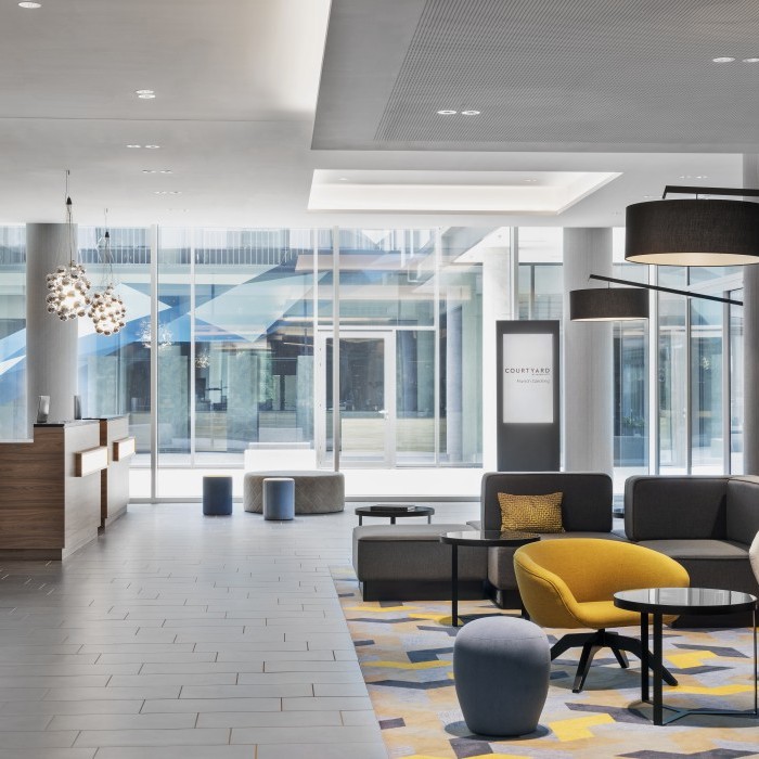 Helle und moderne Lobby in einem Hotel nahe dem Flughafen München mit stilvollen Sitzgelegenheiten in Grau und Gelb, dekorativen Leuchten und großen Glasfenstern im Hintergrund.