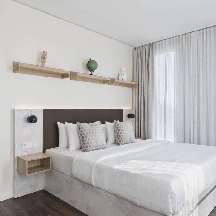 Modernes Zimmer mit einem großen Doppelbett, minimalistischer Dekoration, Holzregalen und bodentiefen Fenstern mit weißen Vorhängen im Apartmenthotel in Garching bei München.