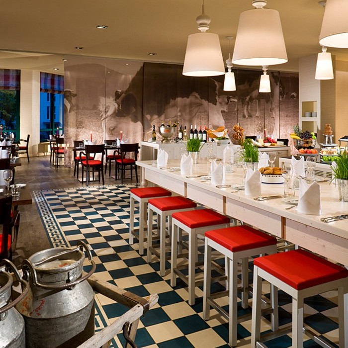 Gemütliches Restaurant mit rustikalem Charme, großen Holztischen, rot gepolsterten Hockern, stilvoll gedeckten Plätzen und ländlichen Dekoelementen im Flughafenhotel München (Freising).