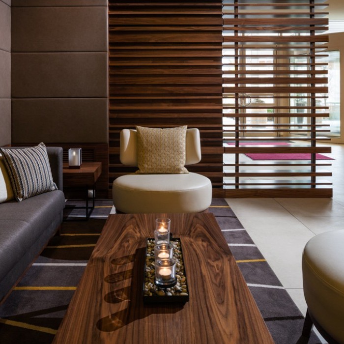 Hotel-Lounge mit modernem Holztisch, bequemen Sitzmöbeln und einer stilvollen Holzwand neben einem gläsernen Eingang im Flughafenhotel München (Freising).