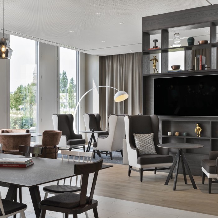 Heller Loungebereich mit gemütlichen Sesseln, großem Tisch, dekorativem Regal mit Fernseher und bodentiefen Fenstern mit Blick ins Grüne im Apartmenthotel in Garching bei München.