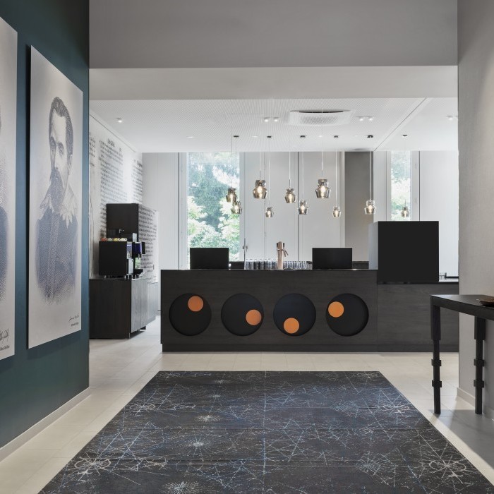 Eleganter Empfangsbereich mit modernen Pendelleuchten, dekorativem Teppich, stilvoller Wandkunst und dunkler Rezeption mit geometrischen Akzenten im Apartmenthotel in Garching bei München.