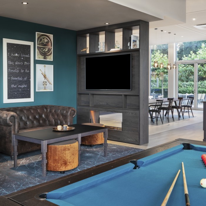 Gemütlicher Freizeitbereich mit Billardtisch, Chesterfield-Sofas, modernen Dekoelementen, Wandkunst und einem großen Fernseher vor einer blauen Akzentwand im Apartmenthotel in Garching bei Mü