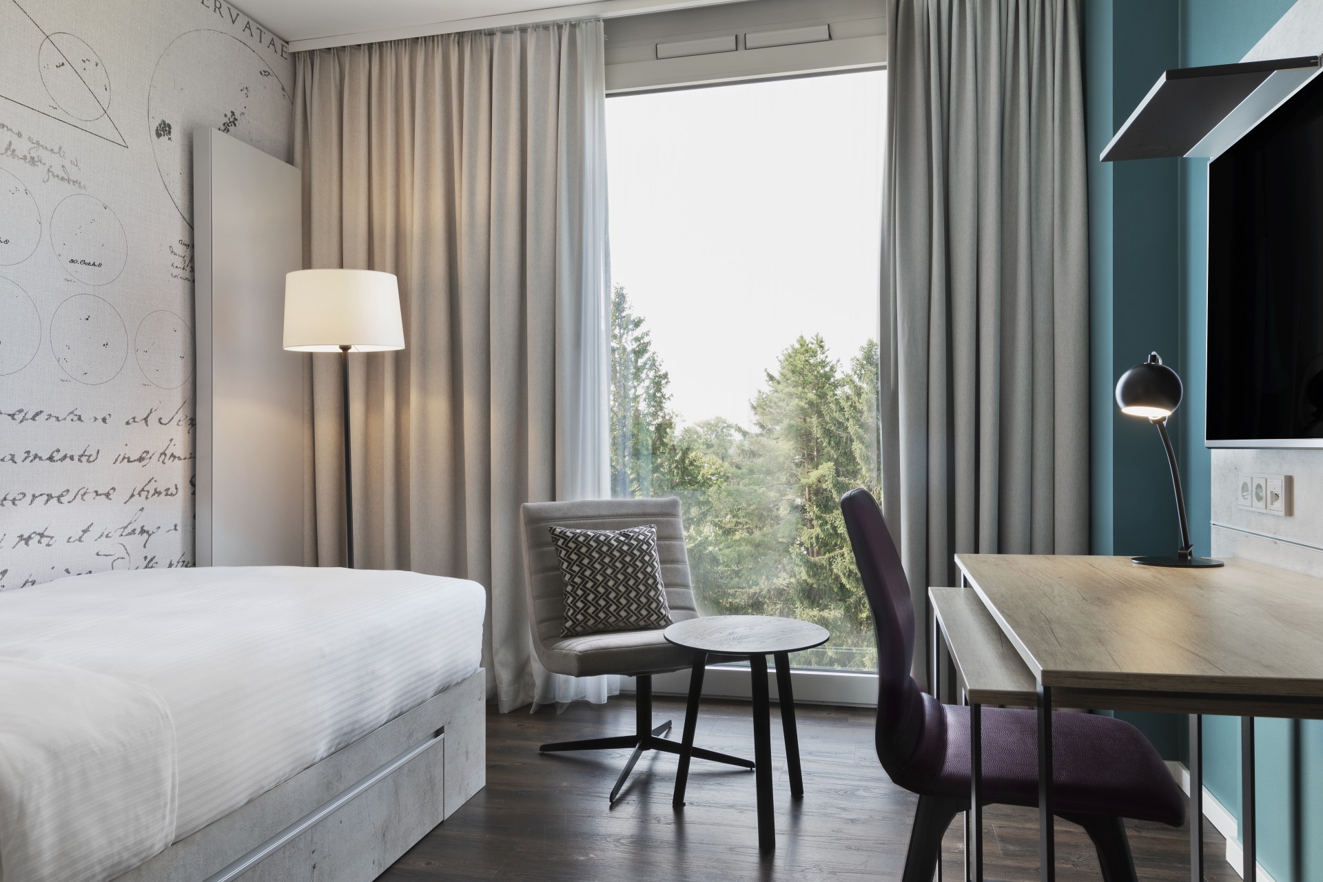 Modernes Zimmer mit schlichtem Design, einem großen Fenster mit Blick ins Grüne, Schreibtisch, gemütlichem Sessel und dekorativer Wandgestaltung im Apartmenthotel in Garching bei München.