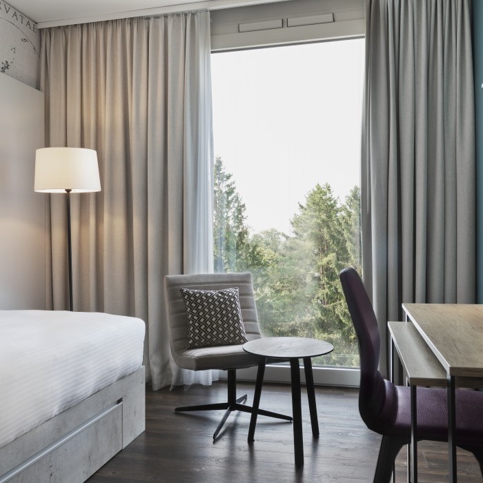 Modernes Zimmer mit schlichtem Design, einem großen Fenster mit Blick ins Grüne, Schreibtisch, gemütlichem Sessel und dekorativer Wandgestaltung im Apartmenthotel in Garching bei München.