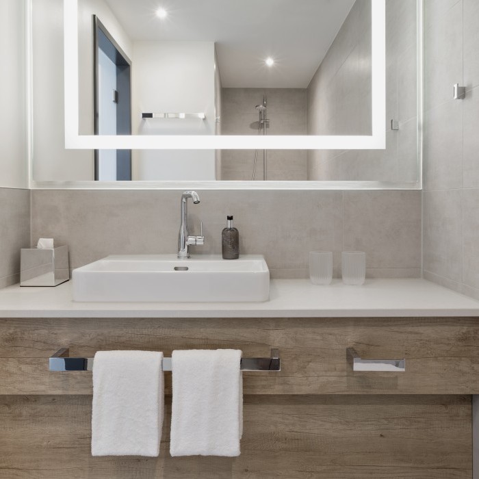 Modernes Badezimmer mit Waschbecken, großem beleuchtetem Spiegel, minimalistischer Einrichtung und hellen Fliesen im Apartmenthotel in Garching bei München.