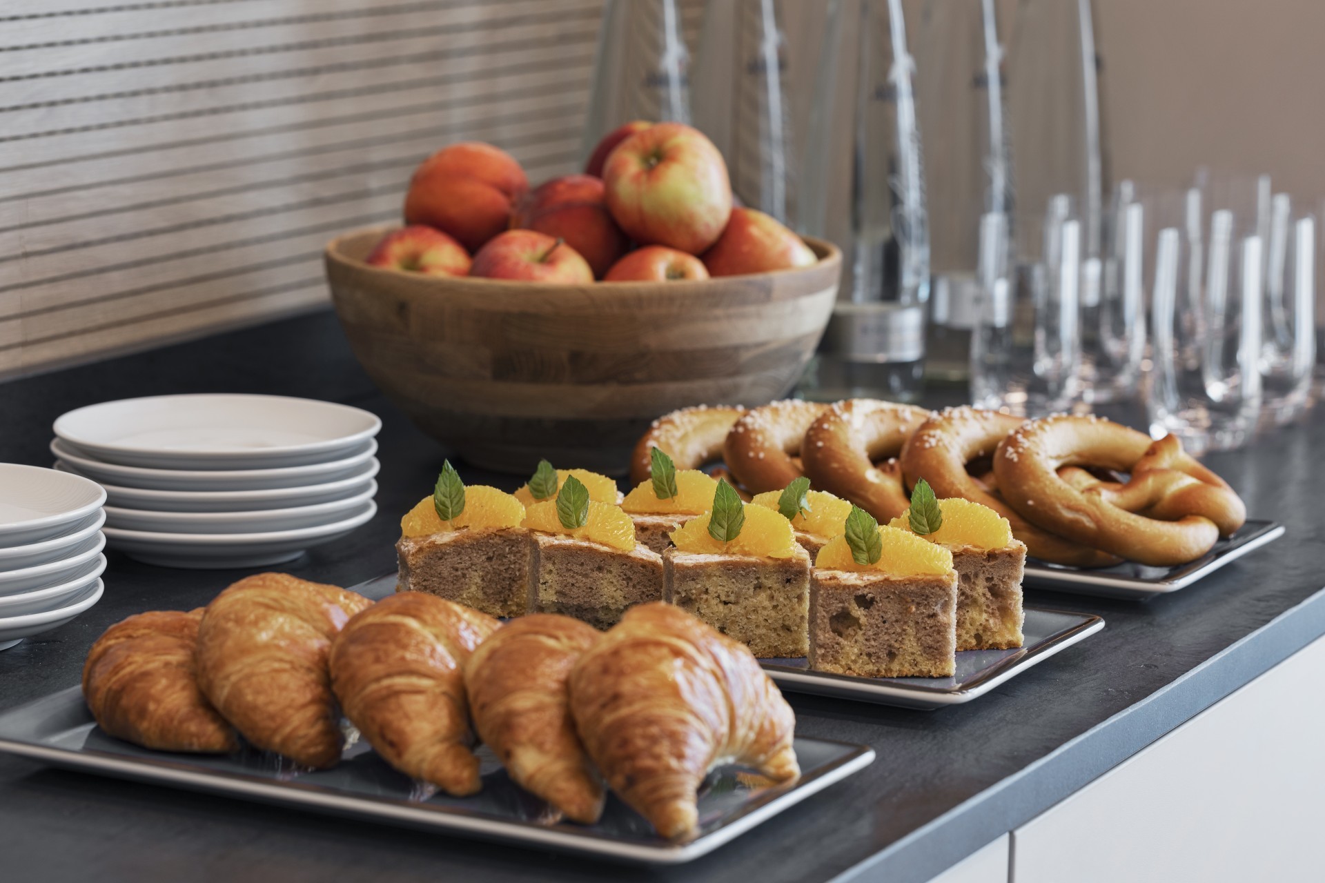 Frühstücksbuffet mit frischen Croissants, Brezeln, Obstkuchen mit Orangenscheiben, einer Schale voller Äpfel und gestapelten weißen Tellern auf einem Tisch.