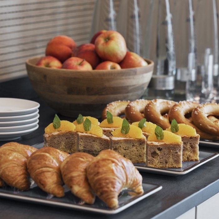 Frühstücksbuffet mit frischen Croissants, Brezeln, Obstkuchen mit Orangenscheiben, einer Schale voller Äpfel und gestapelten weißen Tellern auf einem Tisch.