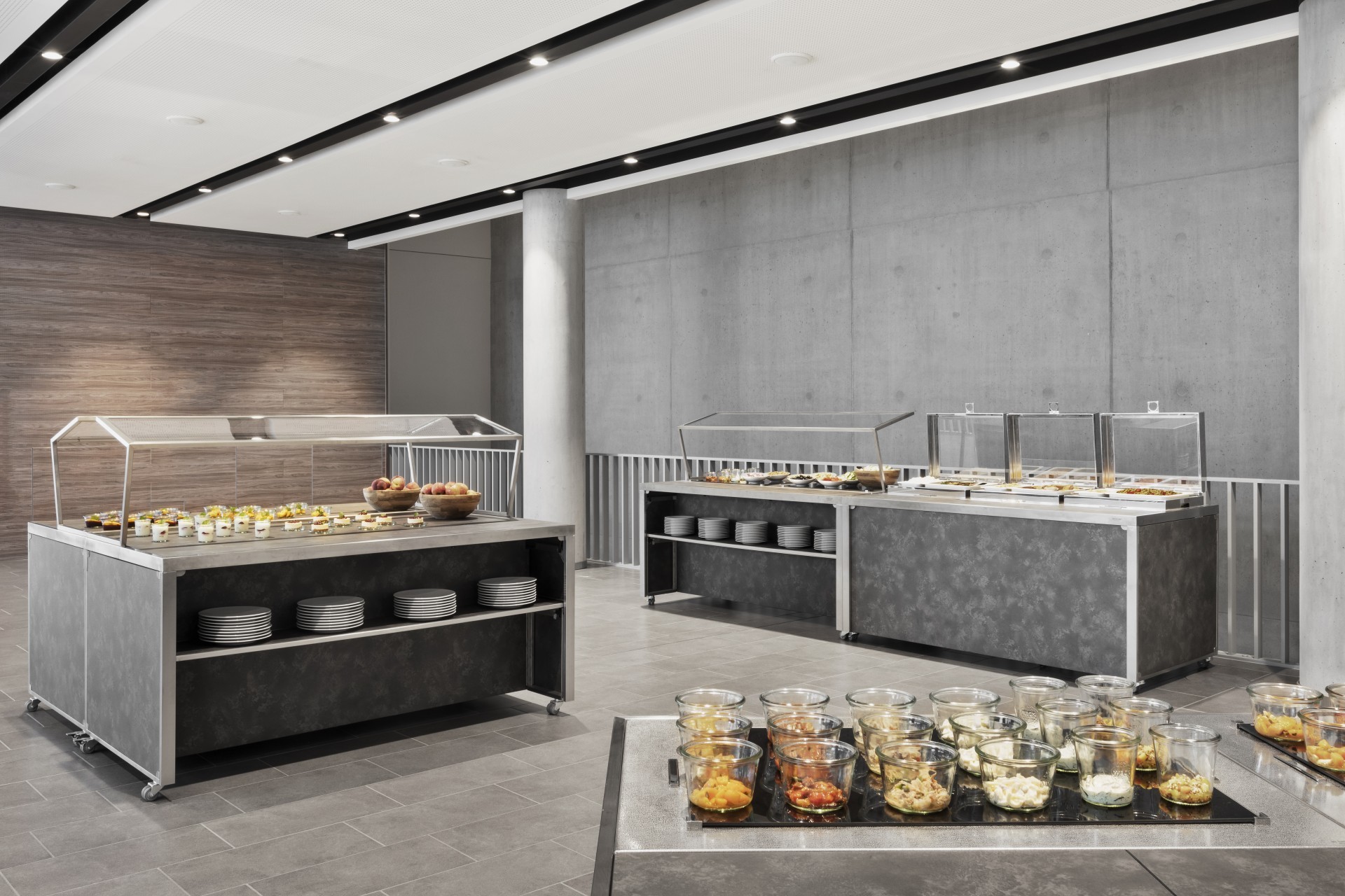 Modernes Buffet mit verschiedenen Speisen in Glasbehältern, warmen Gerichten unter Abdeckungen und gestapelten Tellern in einem stilvollen, minimalistischen Raum.