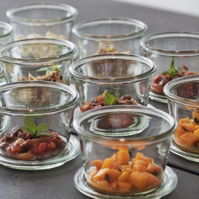 Mehrere Glasbehälter mit verschiedenen frischen Speisen, darunter Gemüse und Salate, dekoriert mit Kräutern auf einer grauen Oberfläche.