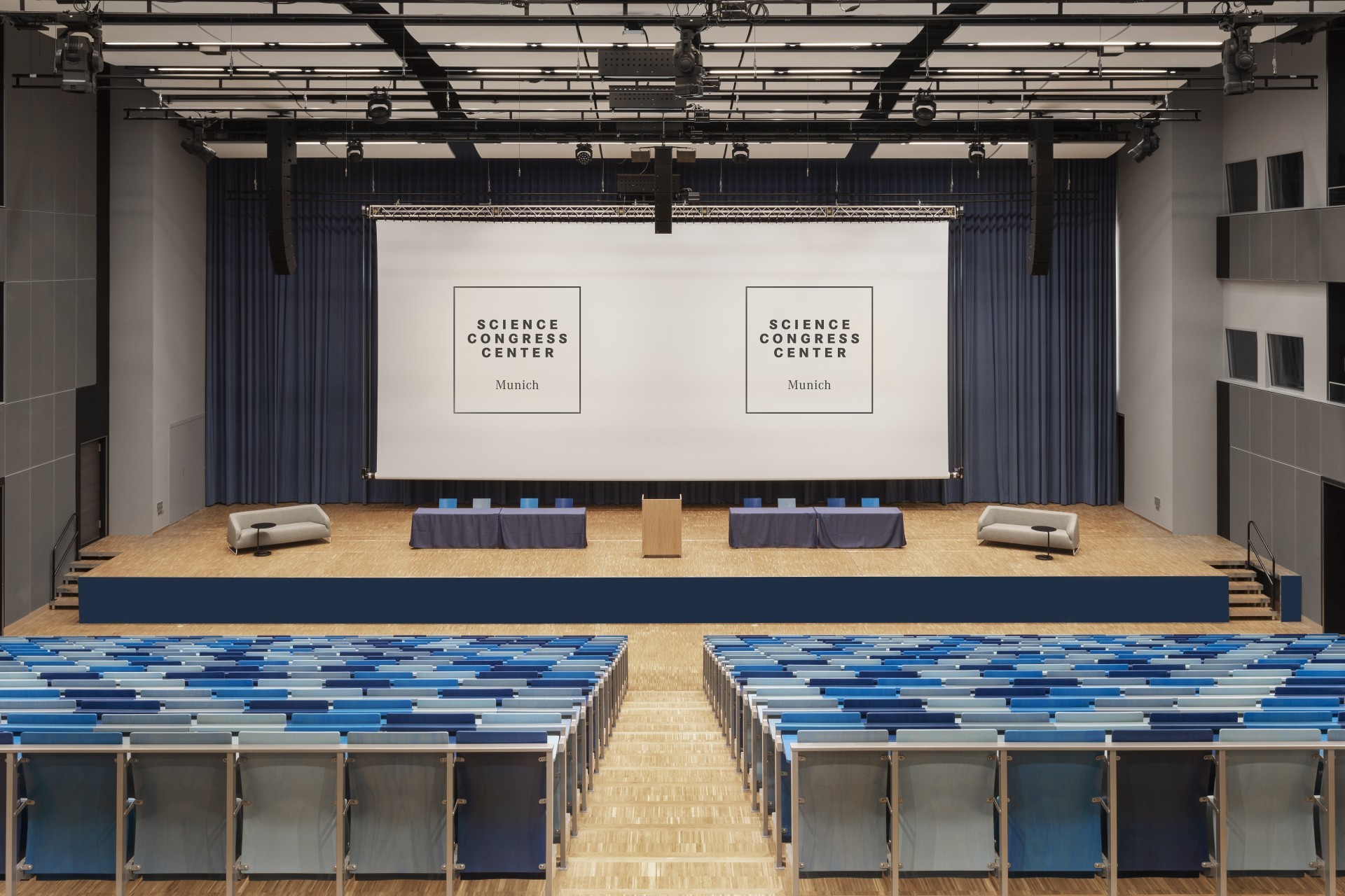 Auditorium im Science Congress Center Munich mit blauen Sitzreihen, einer großen Bühne und zwei Projektionsflächen für Präsentationen.