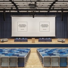 Auditorium im Science Congress Center Munich mit blauen Sitzreihen, einer großen Bühne und zwei Projektionsflächen für Präsentationen.