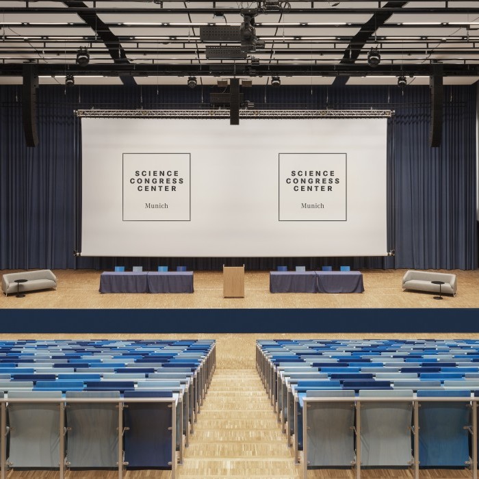 Auditorium im Science Congress Center Munich mit blauen Sitzreihen, einer großen Bühne und zwei Projektionsflächen für Präsentationen.