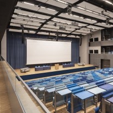 Modernes Auditorium mit blauen Sitzreihen, einer großen Bühne und einer weißen Leinwand, ideal für Präsentationen und Konferenzen.