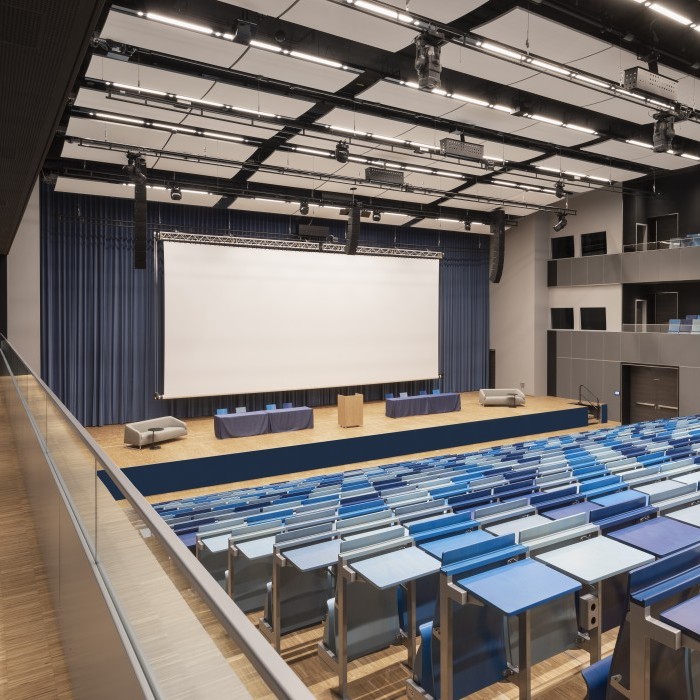 Modernes Auditorium mit blauen Sitzreihen, einer großen Bühne und einer weißen Leinwand, ideal für Präsentationen und Konferenzen.
