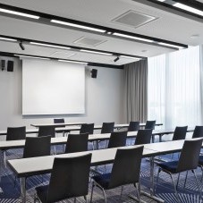 Moderner Seminarraum mit blauen Stühlen, weißen Tischen, einer Leinwand und bodentiefen Fenstern für viel Tageslicht.