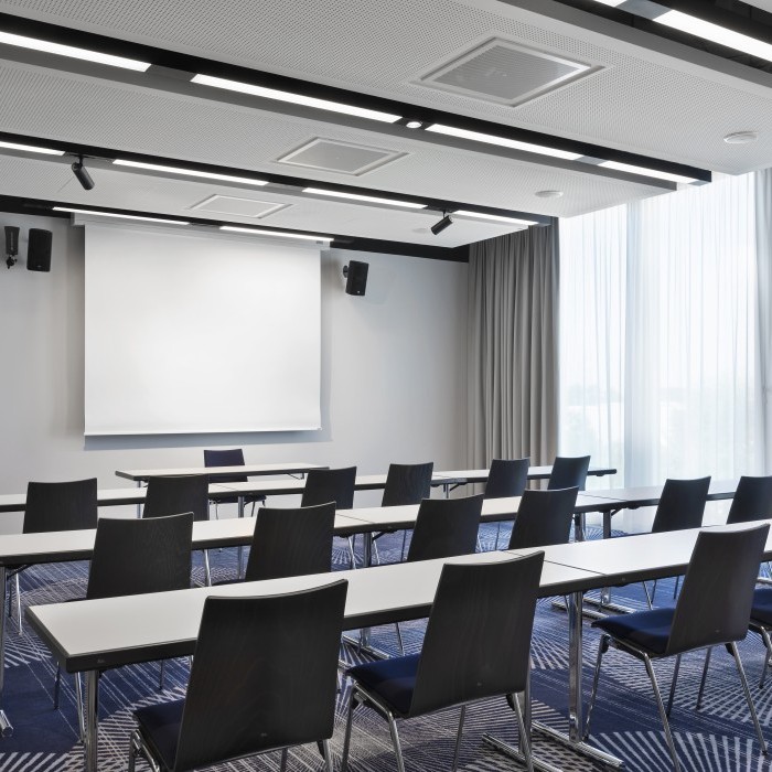 Moderner Seminarraum mit blauen Stühlen, weißen Tischen, einer Leinwand und bodentiefen Fenstern für viel Tageslicht.