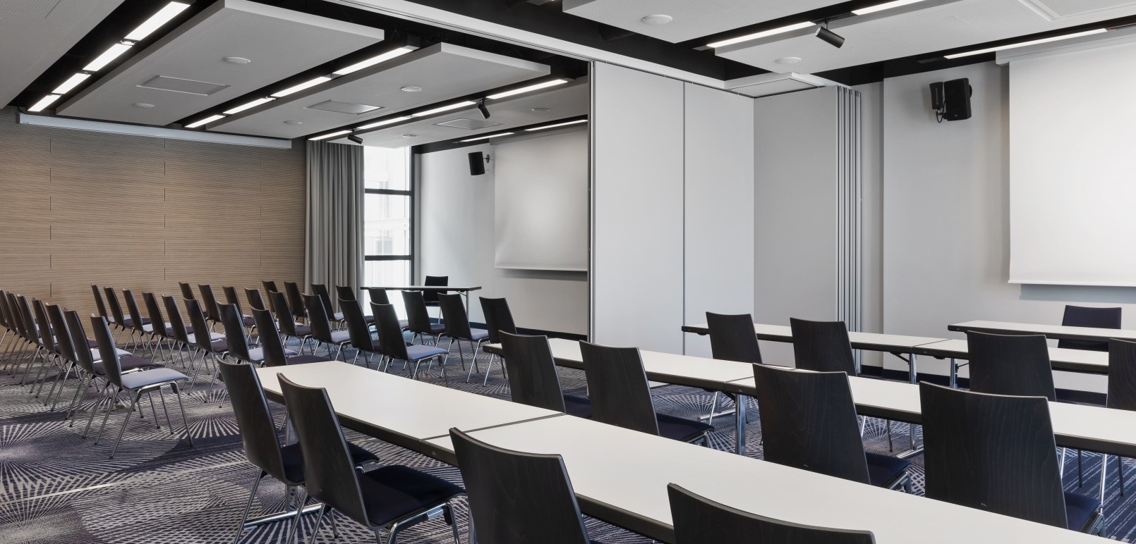 Moderner Konferenzraum mit Reihen aus schwarzen Stühlen, weißen Tischen, Präsentationstechnik und schallabsorbierenden Deckenpaneelen.