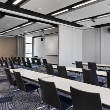 Moderner Konferenzraum mit Reihen aus schwarzen Stühlen, weißen Tischen, Präsentationstechnik und schallabsorbierenden Deckenpaneelen.