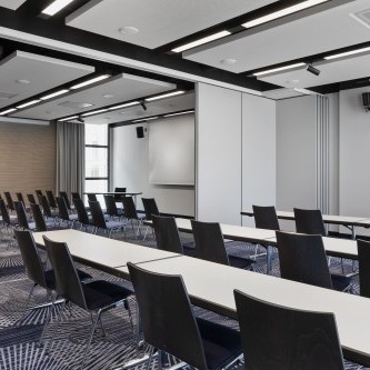 Moderner Konferenzraum mit Reihen aus schwarzen Stühlen, weißen Tischen, Präsentationstechnik und schallabsorbierenden Deckenpaneelen.