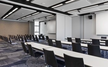 Moderner Konferenzraum mit Reihen aus schwarzen Stühlen, weißen Tischen, Präsentationstechnik und schallabsorbierenden Deckenpaneelen.
