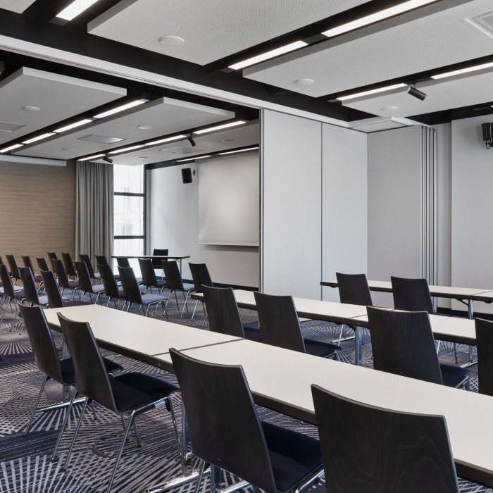 Moderner Konferenzraum mit Reihen aus schwarzen Stühlen, weißen Tischen, Präsentationstechnik und schallabsorbierenden Deckenpaneelen.