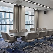 Moderner Besprechungsraum mit großem Konferenztisch, gepolsterten grauen Drehstühlen, bodentiefen Fenstern und stilvollem Teppichdesign.