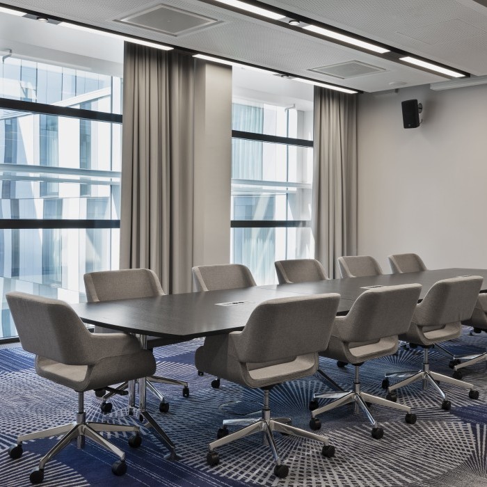 Moderner Besprechungsraum mit großem Konferenztisch, gepolsterten grauen Drehstühlen, bodentiefen Fenstern und stilvollem Teppichdesign.
