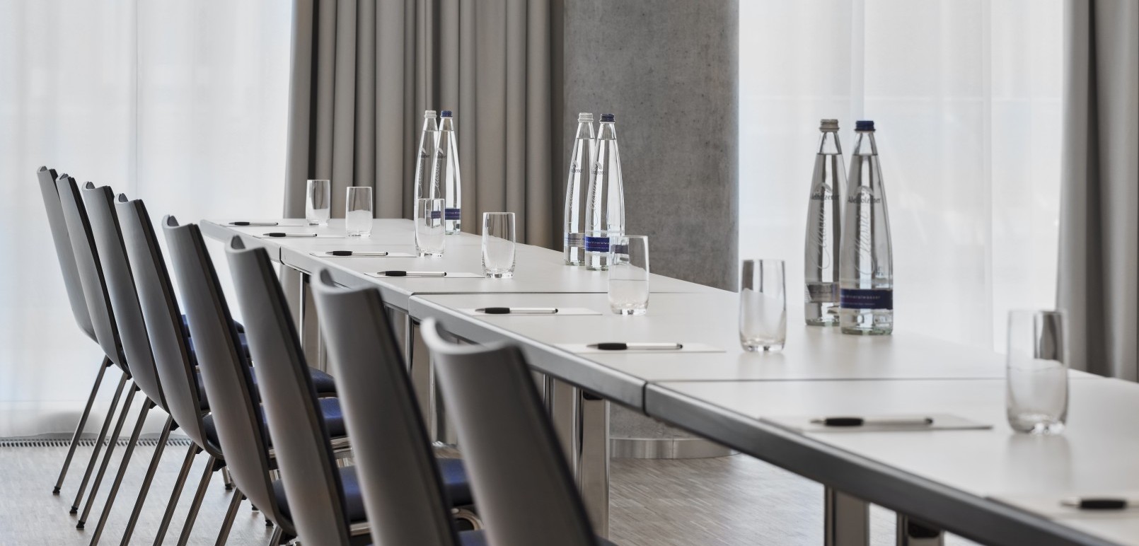 Detailaufnahme eines Konferenztisches mit Stühlen, bereitgestellten Wasserflaschen, Gläsern und Schreibutensilien vor einem Hintergrund aus hellen Vorhängen.