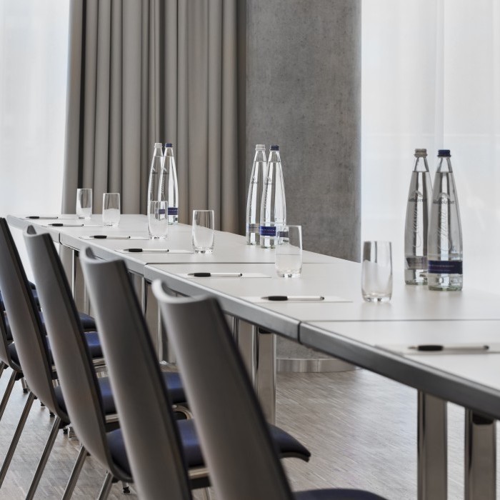 Detailaufnahme eines Konferenztisches mit Stühlen, bereitgestellten Wasserflaschen, Gläsern und Schreibutensilien vor einem Hintergrund aus hellen Vorhängen.