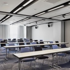 Heller Konferenzraum mit weißen Tischen, farblich gemischten Stühlen, moderner Beleuchtung und einem stilvollen Teppich mit geometrischem Muster.