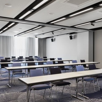 Heller Konferenzraum mit weißen Tischen, farblich gemischten Stühlen, moderner Beleuchtung und einem stilvollen Teppich mit geometrischem Muster.