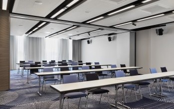 Heller Konferenzraum mit weißen Tischen, farblich gemischten Stühlen, moderner Beleuchtung und einem stilvollen Teppich mit geometrischem Muster.