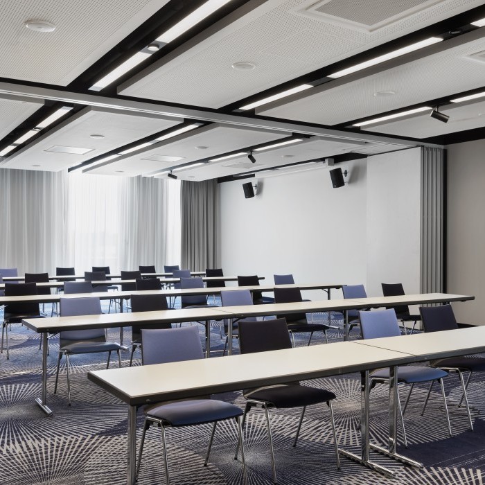 Heller Konferenzraum mit weißen Tischen, farblich gemischten Stühlen, moderner Beleuchtung und einem stilvollen Teppich mit geometrischem Muster.