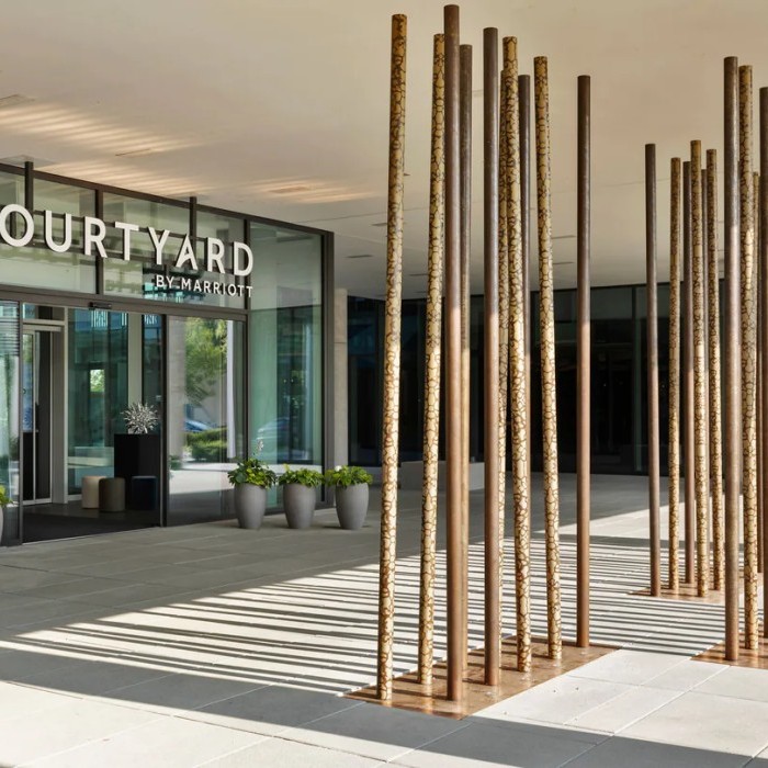 Eingangsbereich des Courtyard by Marriott Hotels mit modernen Holzskulpturen und großen Pflanzenkübeln auf einer gepflasterten Terrasse.