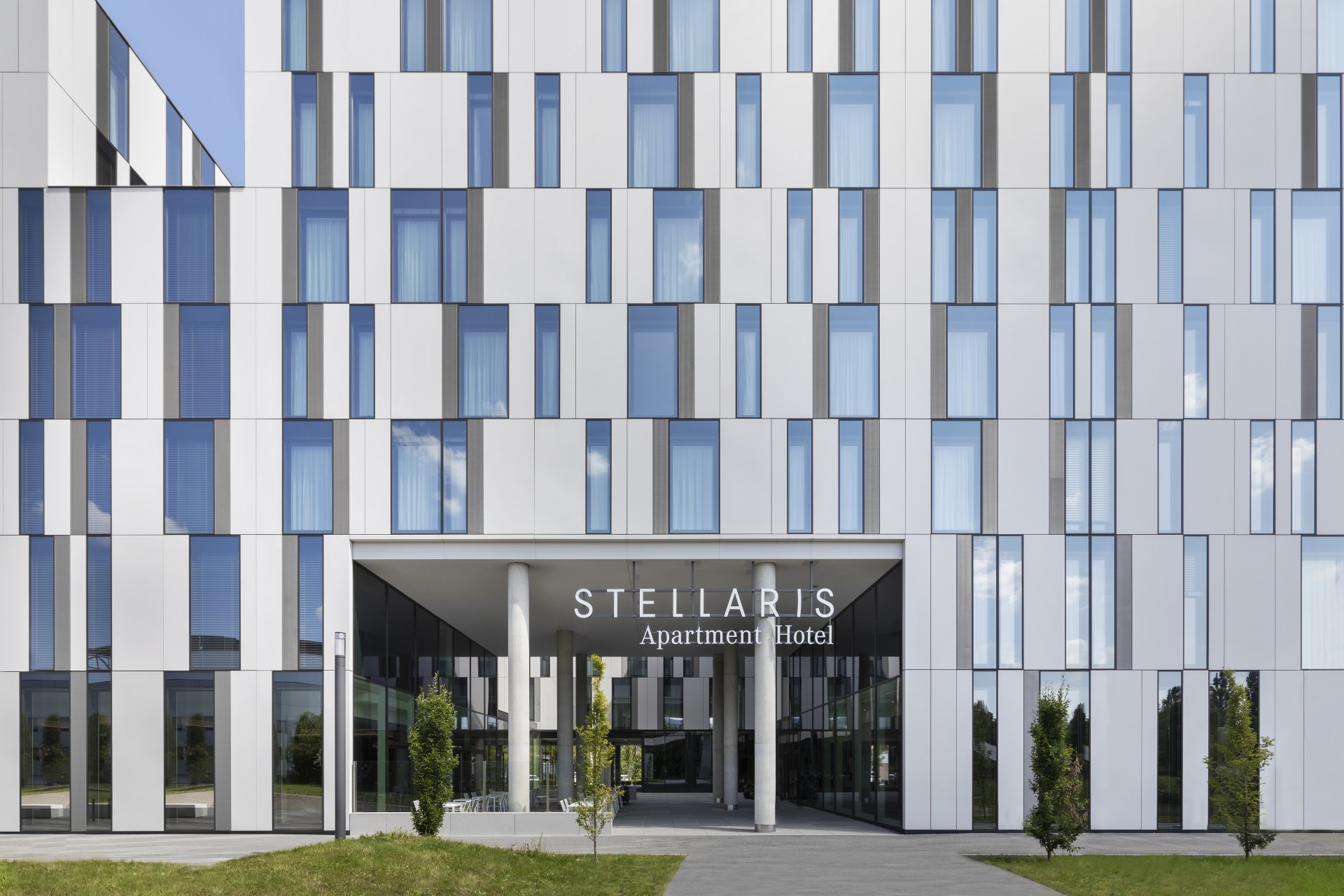 Eingangsbereich des Stellaris Apartment Hotels mit moderner Glasfassade und geometrischem Design, eingerahmt von schlichten Säulen und grünen Pflanzen.