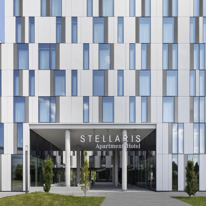 Eingangsbereich des Stellaris Apartment Hotels mit moderner Glasfassade und geometrischem Design, eingerahmt von schlichten Säulen und grünen Pflanzen.